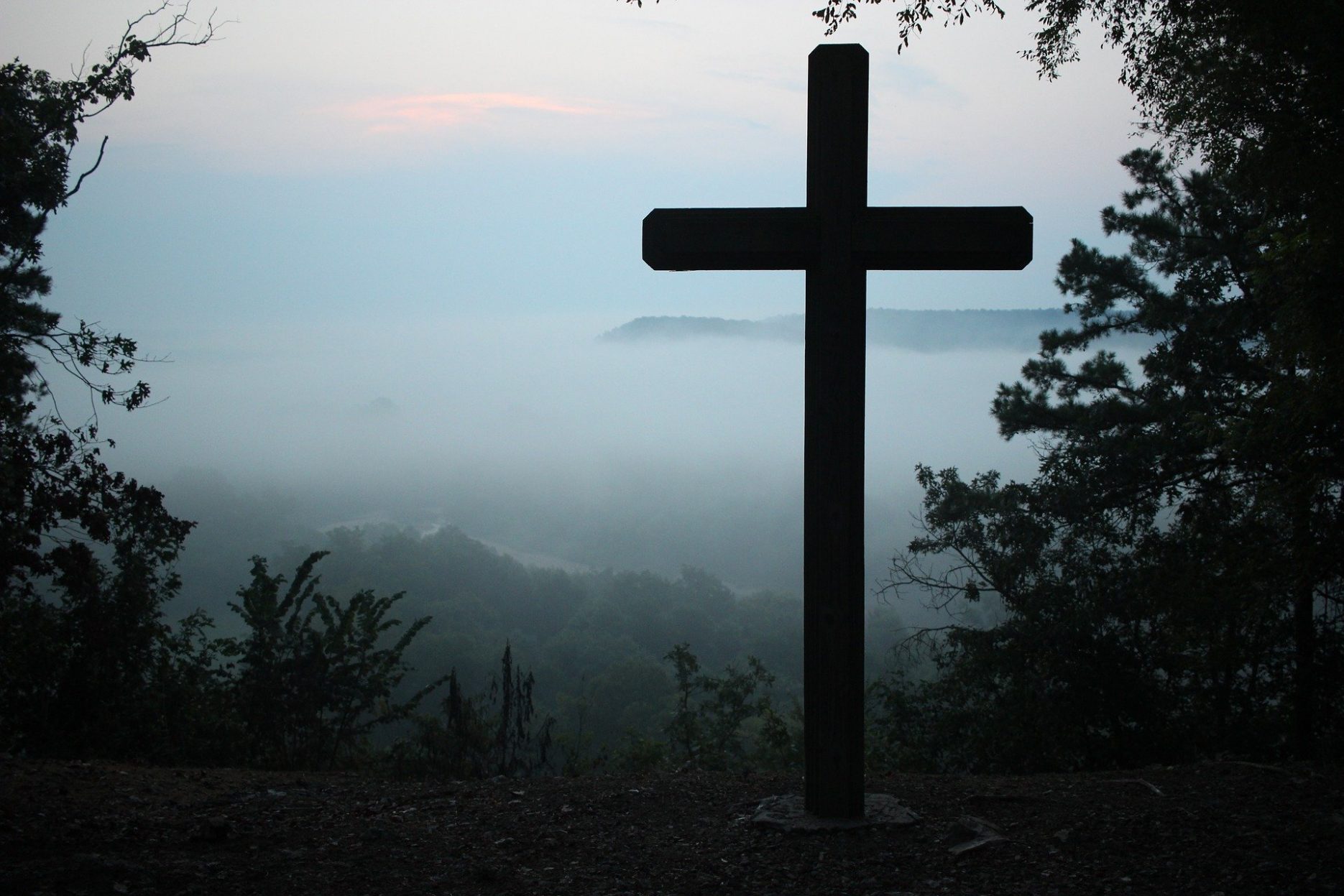 Das Bild zeigt ein großes Kreuz, das auf einem bewaldeten Berg aufgestellt ist. Im Hintergrund liegt das von Nebel bedeckte Tal.