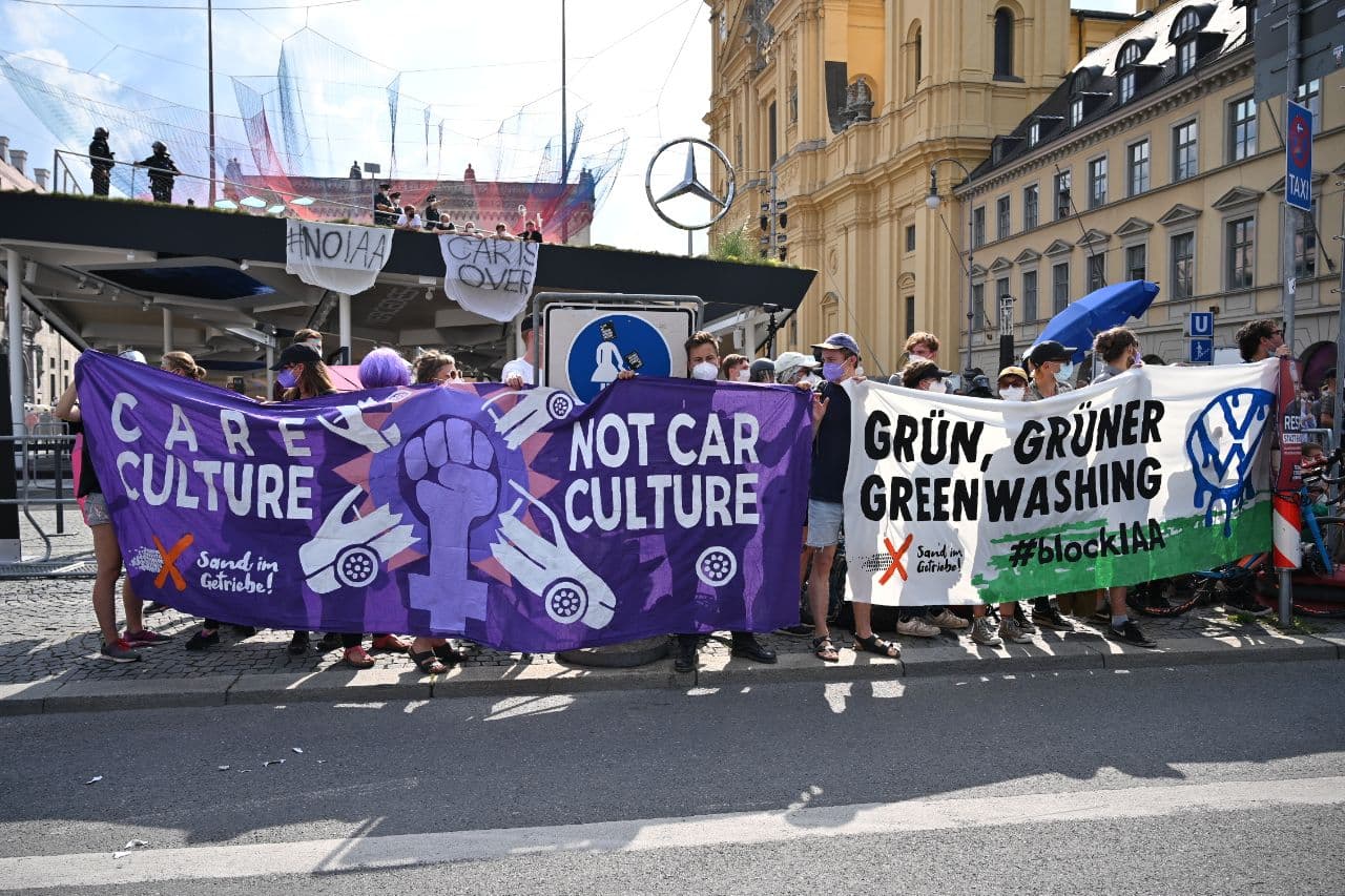 Das Bild zeigt junge Aktivist*innen mit Bannern bei der Internationalen Automobil Ausstellung in der Münchener Innenstadt. Auf den Bannern stehen die Slogans "Care Culture - Not Car Culture" sowie "Grün, Grüner, Greenwashing".