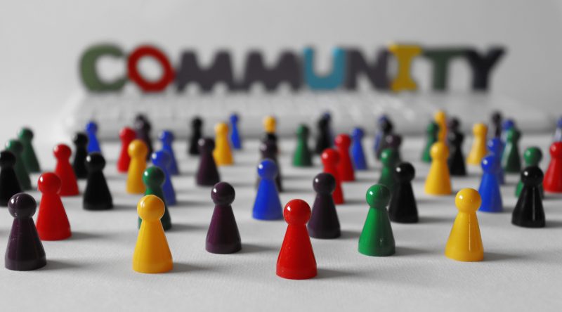 Das Bild zeigt viele verschieden farbige Mensch-ärgere-dich-nicht-Spielfiguren, im Vorder und Hintergrund. Ganz im Hintergrund steht das Wort COMMUNITY geschrieben.