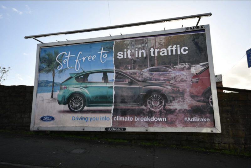 Das Bild zeigt eine Werbetafel mit Plakat der Badvertisment-Kampagne. Dieses zeigt einen SUV, der zur Hälfte mit positiven Assoziationen (Strand, Palmen) und zur Hälfte mit negativen Assoziationen (Stau, Luftverschmutzung) abgebildet ist. Dazu steht geschrieben: "Mach dich frei, um im Verkehr festzustecken". Und: "Fährt dich in den Klimakollaps".