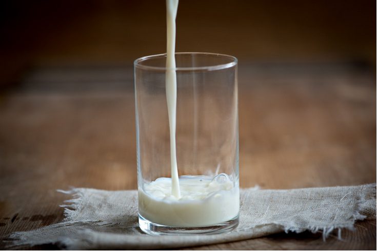 Das Bild zeigt ein Glas auf einem Holztisch und einer kleinen Tischdecke, in das Milch eingegossen wird.