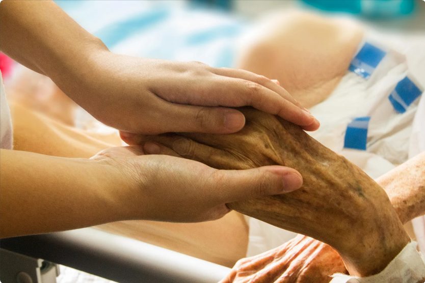 Das Foto zeigt die Hände einer jüngeren Person, die eine Hand einer deutlich älteren Person in einem Krankenhausbett hält.