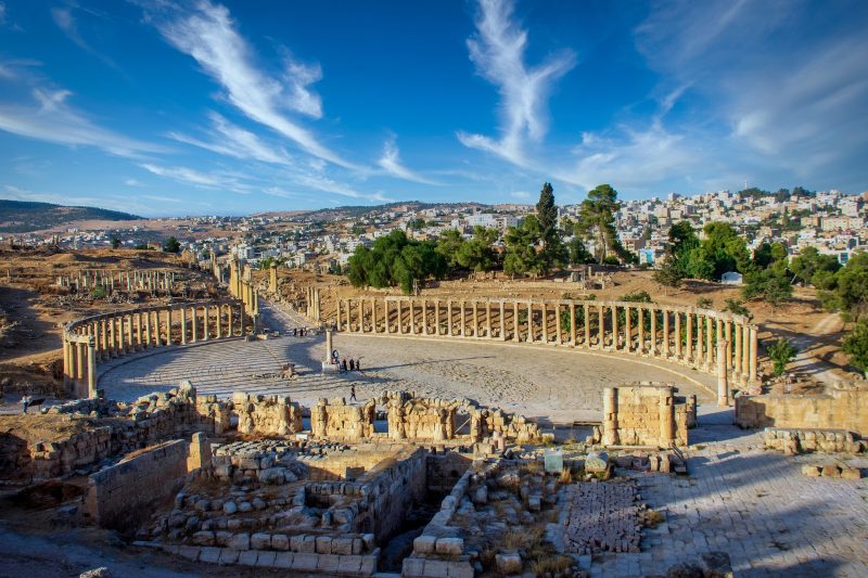 Das Bild zeigt im Vordergrund die Ruinen des antiken Forums der Stadt Gerasa in Jordanien, im Hintergrund die (moderne) Stadt.