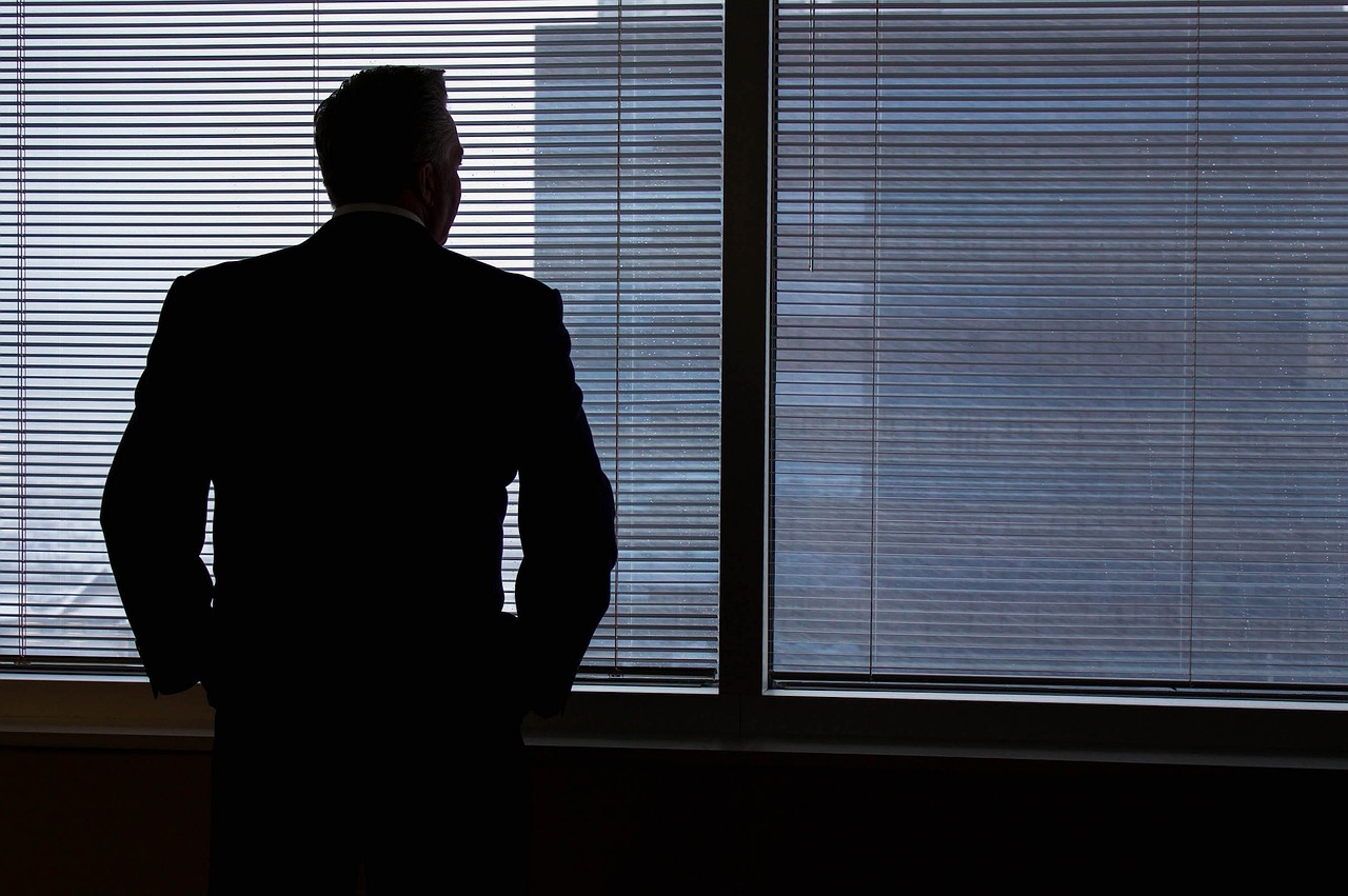 Bild zeigt eine männlich gelesene Person im Anzug, die aus dem Fenster eines hohen Bürogebäudes blickt.